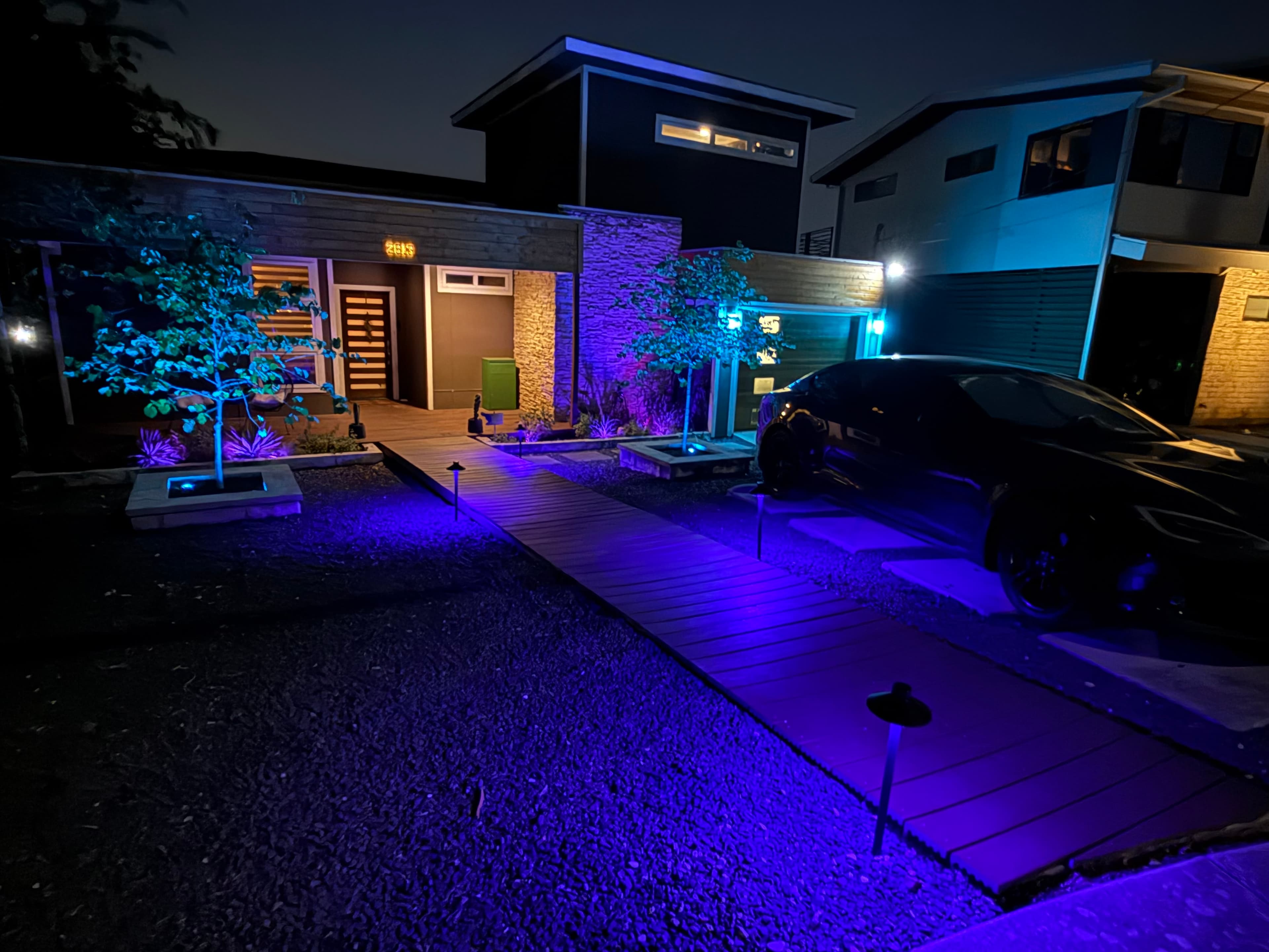 Modern house at night with vibrant purple and blue landscape lighting and wooden walkway.